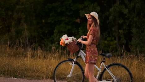 Side-view-of-smiling-sexy-blonde-girl-in-a-light-summer-dress-is-walking-with-a-Bicycle-in-the-summer-in-the-handlebar-basket-of-wildflowers-in-slow-motion